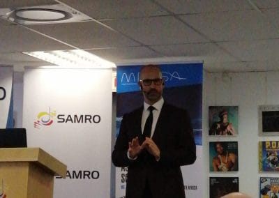 A man in a dark suit, white shirt, and glasses stands speaking in front of a podium with a “SAMRO” sign. Behind him are banners, posters, and several seated people, with visible heads in the audience. The event appears to be held indoors under ceiling lights.