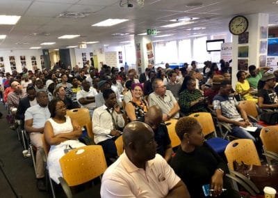 A diverse group of people sit in rows of chairs, attentively facing forward in a well-lit room. The audience includes men and women of various ages and backgrounds. Walls display photos and posters; a clock and windows are visible in the bright space.