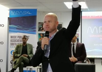 A bald man in a suit stands foreground, holding a microphone and raising his left hand while speaking. Behind him, two men sit on chairs, one in a green suit and the other in a dark suit and glasses. Music industry banners and a screen are visible in the background.