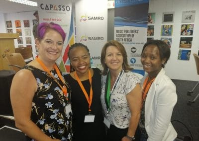 Four women smile together at a professional event, each wearing orange lanyards. They stand in front of banners for CAPASSO, SAMRO, and IMPA SA, related to music publishing in South Africa. Background shows a decorated room with posters and chairs.