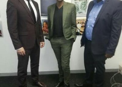 Three people stand indoors in front of a wall decorated with various album covers. Two men wear dark suits with dress shoes and shirts, while the person in the center wears a green suit, black shirt, and glasses. All are smiling and facing the camera.