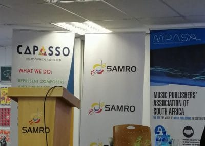 A conference setup with a wooden podium, two chairs, a small table with water bottles, and banners for CAPASSO, SAMRO, and MPASA. The banners promote music rights organizations in South Africa. The room has a white ceiling with fluorescent lights.
